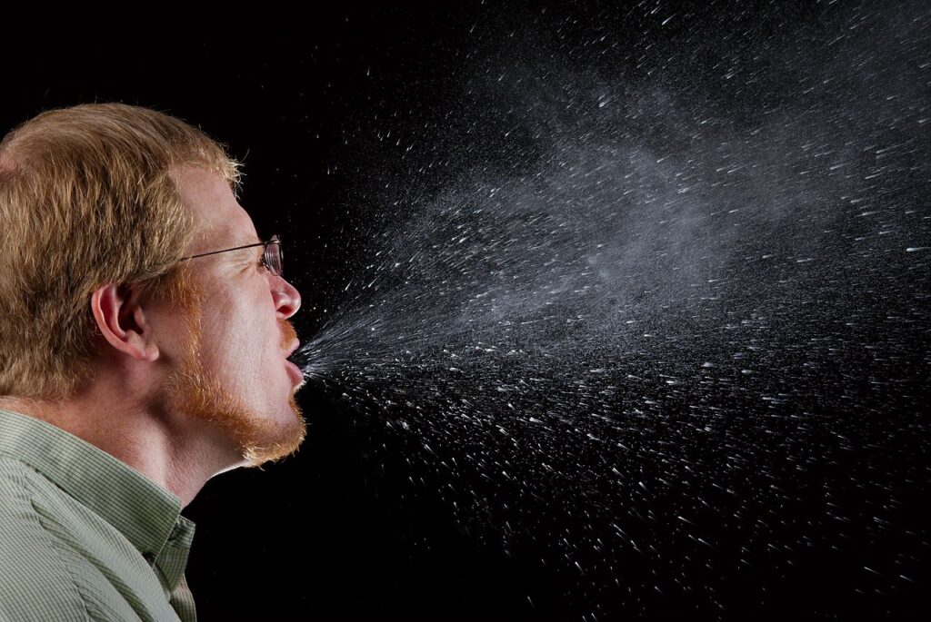 High-speed camera captures bright droplets flying from a person’s uncovered cough versus minimal spray when wearing a surgical mask.
